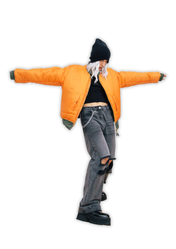 Model wearing an orange bomber jacket, black crop top, and ripped grey jeans with a chain accessory, posing in a fashionable urban style.